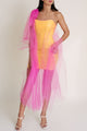 Woman is posing and wearing a yellow dress with a sheer pink overlay and white heels.