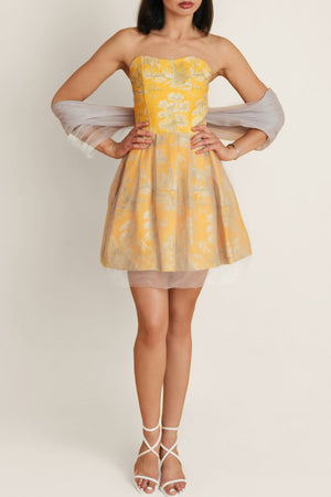 Woman in golden yellow Porto Chic cotton and tulle dress with yellow-gray print, paired with white shoes.